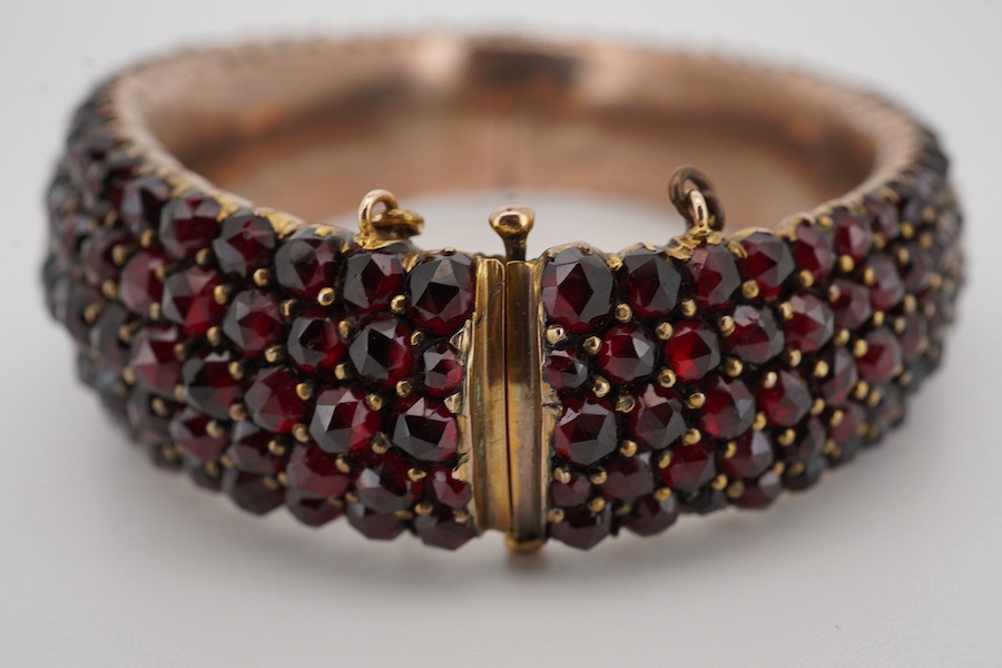 An Edwardian yellow metal overlaid and facetted garnet cluster set hinged bracelet, with safety chain, interior diameter 59mm. Condition - poor to fair